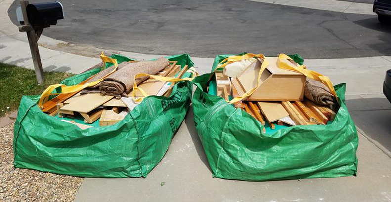 Two Bagster bags next to each other at the end of a driveway filled with carpet rolls, wood material, and cardboard.